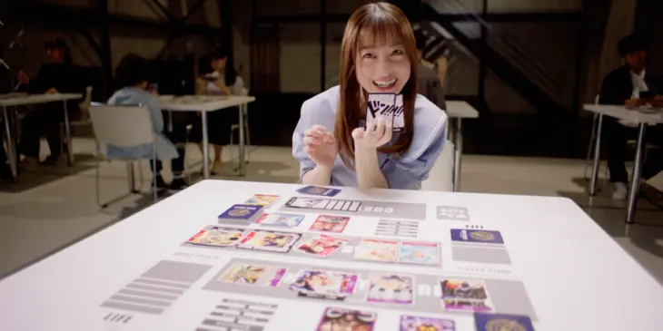 Kanna Hashimoto segura cartas diante de uma mesa de One Piece Card Game em cena promocional da Bandai