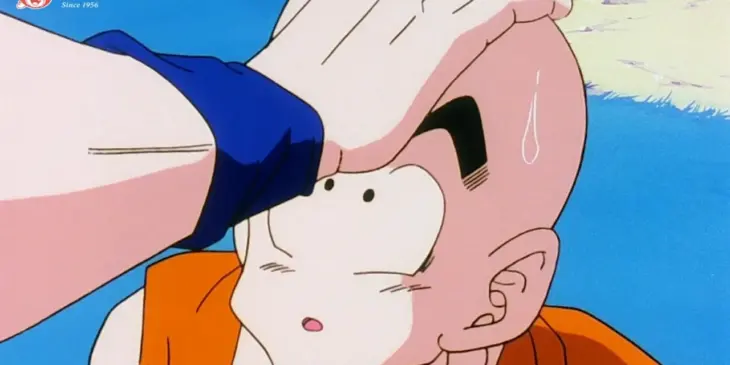 Kuririn de uniforme laranja recebe uma mão sobre a cabeça em uma cena de Dragon Ball